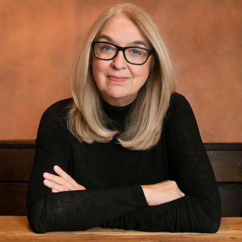 Author Ann Hood, woman with long blond hair wearing dark eyeglasses and a black turtleneck sweater. Her arms are crossed on a wooden table